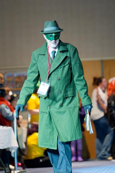 Man in blue pants, green double breasted jacket, green mask and green hat loots at me suspiciously