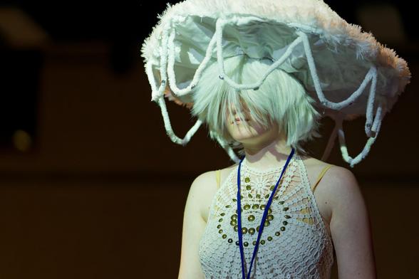 Person wearing mesh blouse with exposed shoulders with circles of circles,  a light greenish wig obscuring the face,  and a fuzzy hat with ropes hanging around it