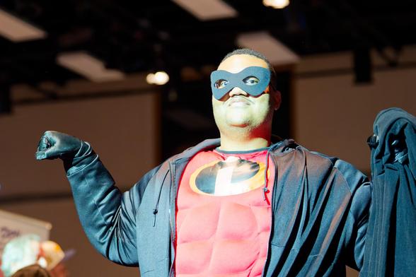 Black man wearing red costume with muscle panels and an i logo in an ellipse and a black mask and jacket