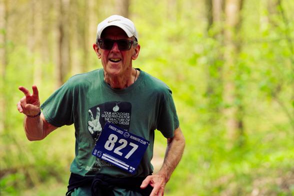 Older man with sunglasses and baseball cap and green shirt makes V hand gesture in the woods
