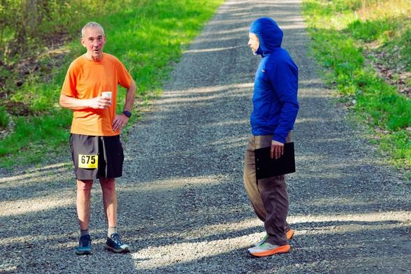 Runner in orange shirt holds cup of water as he talks to man in blue hoodie