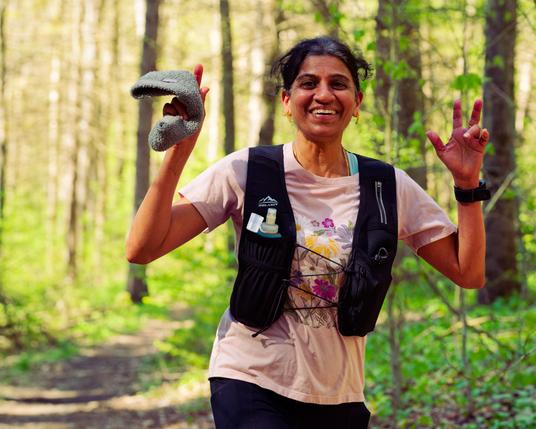 South asian woman with peach shirt holds up hands and poses in the woods