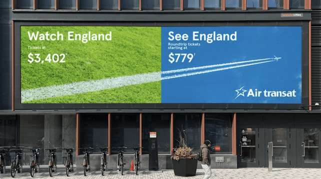 Billboard on the side of a building is divided into two halves.
On the left side over a patch of green grass are the words "Watch England: tickets at $3,402"
On the right side over a blue sky are the words "See England: roundtrip tickets starting at $779"
The white field-marking line on the left side becomes an airplane's trail on the right side.