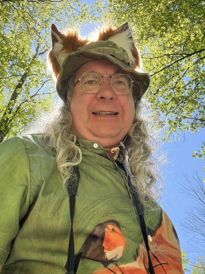 Man with grey hair has a green camoflauge hat with orange/white fox ears on top -- the sun is behind him and all that floof is backlit!  he's got a green shirt with a robin balanced on the nose of a fox,  grey drill hair and the straps of a camerfa.