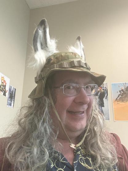 Foxographer with grey ears with white floof and green camo boonie hat and glasses and lots of curly hair with a dark blue shirt with yellow lines and a maroon jacket.  Behind him is a corner you've seen in many selfies I post with lots of anime and mobile game images but not enough contrast