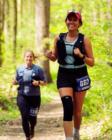 Closer and to the right is as girl with darker than average skin and red cap with glasses on top and further away and to the left is another woman with blond hair and behind lots of tree trunks and leafed out bushes