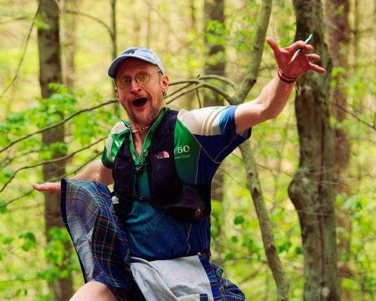 Man with green white and blue shir spreads arms wide and opens his mouth wearing light blue cap in the woods.