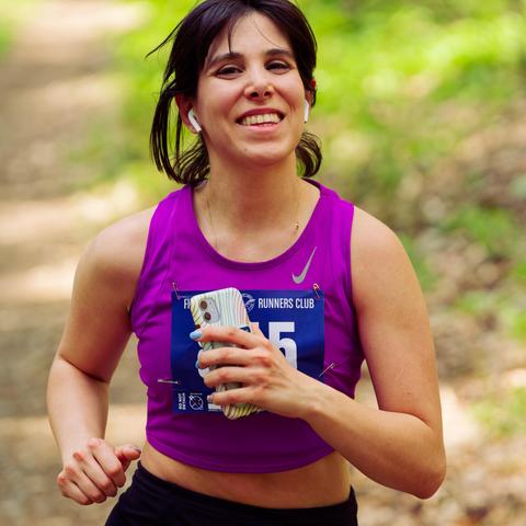 Woman with moderate length black hair has purple tank top with open midriff and black pants and is holding a phone as she runs down a trail.