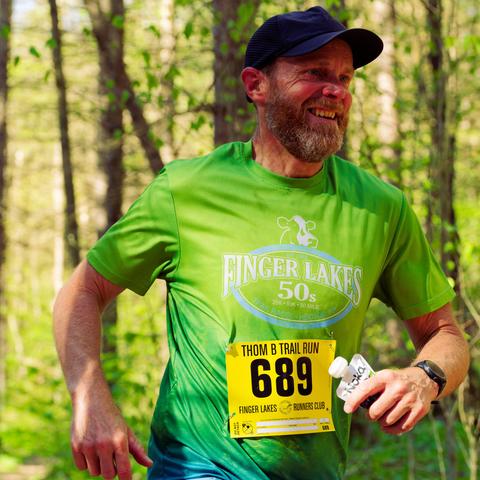 Man in green shirt that says finger lakes 50 has a beard and a blue baseball cap and is holding some kind of food or drink pouch in the woods