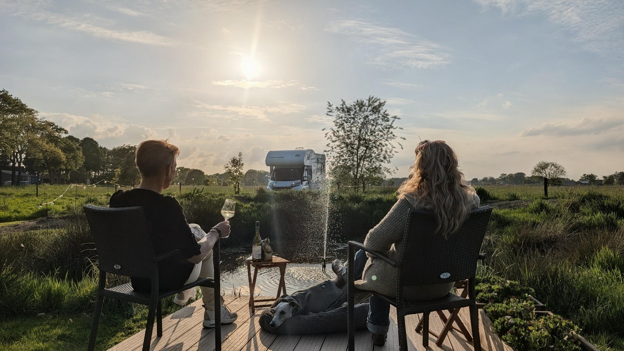 Two people sitting on outdoor chairs on a wooden deck, enjoying wine and the sunset, with a camper van and a small water fountain in the background, surrounded by greenery.