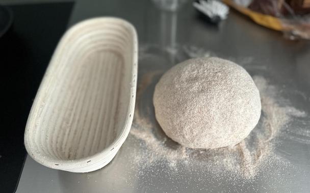 Ein längliches Gärkörbchen aus Bast steht auf der Anrichte. Daneben eine gut bemehlte Teigkugel.