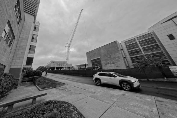 SUV looks small in a road in front of me but the sidewalk in front looks huge although buildings curve in fron the left and white crowding a big crane under a moody overcast sky