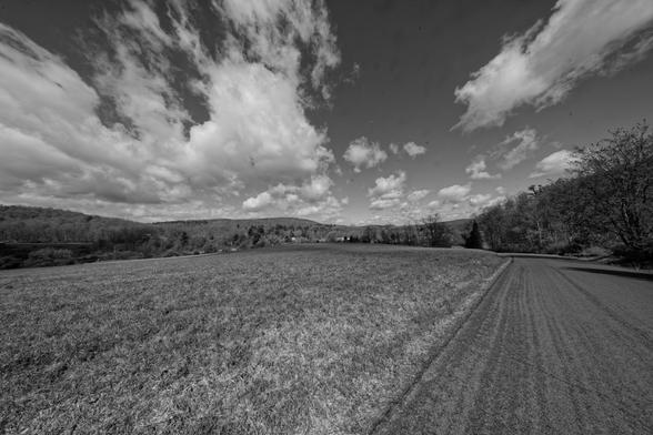 Black and white photo of stretched out cumulus clouds in the sky over a ridge of hills  with road emerging from the lower right corner to proceed down a hill with most of the field to left a field of stubble