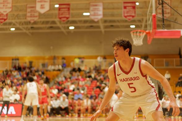 Man crouches in lower right corner with white jersye with red lettering and trim that says HARVARD 5 with blurred out bleachers and net and pennants on the ceiling looming.
