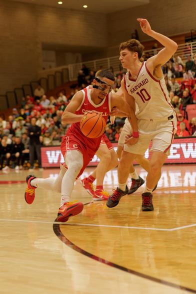 Man with funny plastic facemask dribbles the basketball in the center in a red jersey while number 10 from harvard in a white jersey tries to muscle in