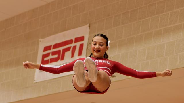 Woman with hair tied close to her face is falling with her arms out in front of a cinderblock wall with an ESPN banner