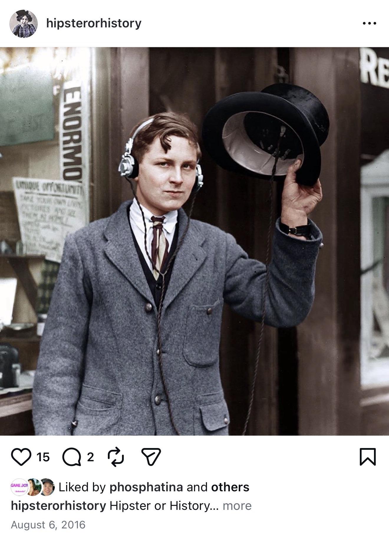 A young man stands wearing headphones and holding a top hat. He is dressed in a gray coat and cravat, with a serious expression. A shop window behind him features a sign, and the image appears to be a vintage photo that has been color