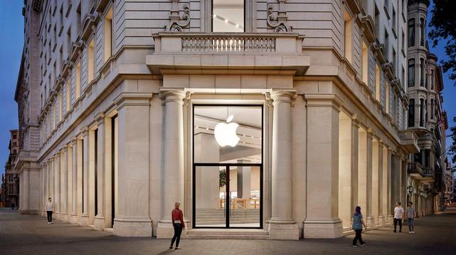 n exterior view of a storefront on the ground floor of a large, light-colo light-colored stone building at a street corner. The building features prominent columns, ornate masonry, and a large glass storefront. A bright white Apple logo is visible through the large glass window. Several people are walking on the sidewalk in front of the building. The scene is captured during twilight, with the edges of adjacent buildings visible against a dark sky.