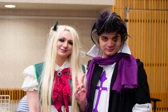 On the left a woman with long blonde hair and a green and white top with red bow making a V and on the right a man with a black coat and white shirt with a purple cross and scarf who looks like Prince to me.