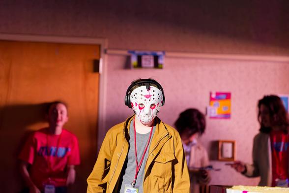 Man wears white mask with numerous small holes and red marks on the forehead and near the eyes with a brown jacket and grey shirt,  behind the wall has a peach cast and you can see a few conference organizsers behind.