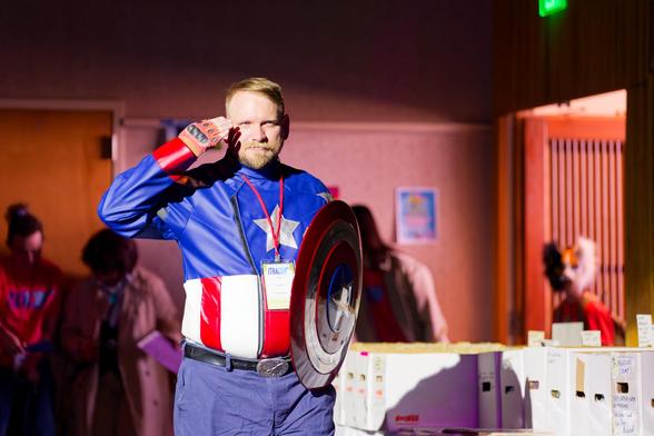 A man with a round shield with star roundel and a shirt with a blue field with one star on top and vertical red and white stripes below salutes next to white boxes full of comics while somebody in the background looks on with a therian mask