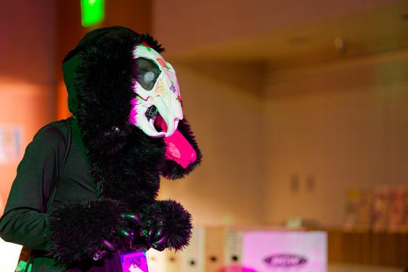 Person wearing dark grey jacket with a lot of black fur around the hood,  a skull painted with spots of color and furry claw gloves
