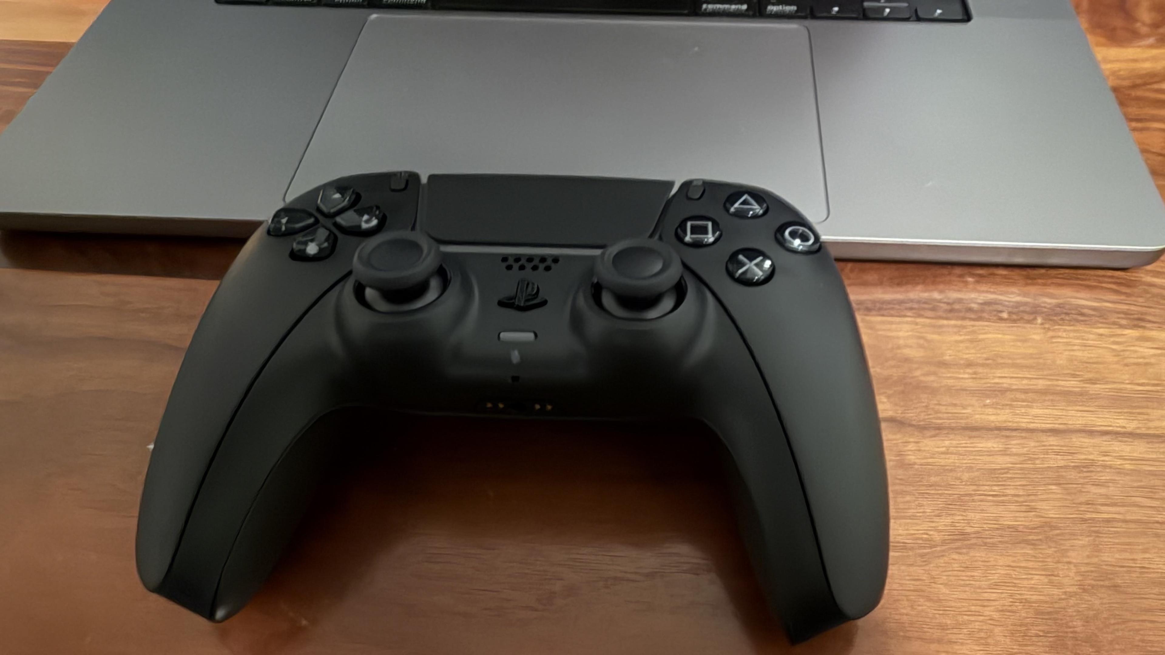A black PlayStation gaming controller is placed on a wooden desk, next to a silver laptop. The controller features two analog sticks and four main buttons.