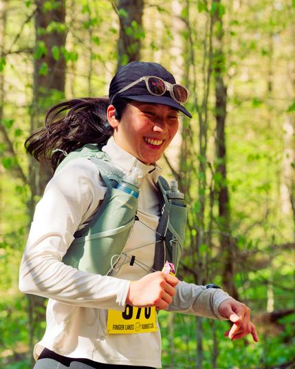 Asian woman with white blouse green vest and blue gap with trees behind.