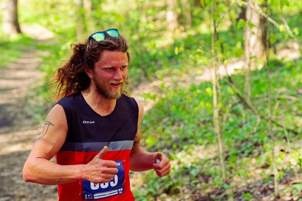 Male runner with long hair and mirrorshades has a blue and red tank top as he runs on trail past understory vegetation