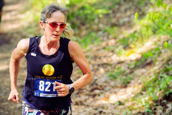 Woman in blue tank top with silver hair, amber shades and muscular arms runs down barren track with a bit of grass to the right side