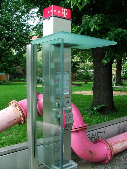 A telephone box with a magenta-pink Deutsche Telekom logo, and a handset in the same colour. Behind it are large drainage pipes connected together in the same magenta-pink colour.