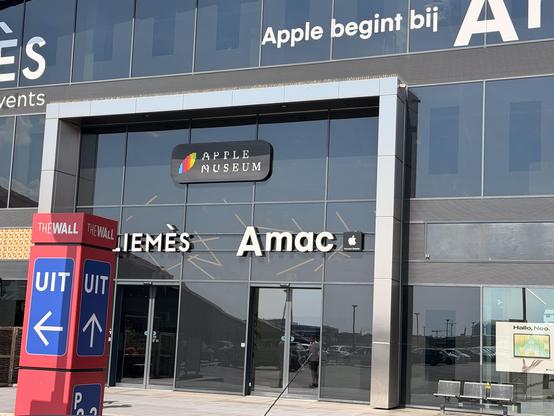 Apple Museum und Amac Store in einem modernen Gebäude mit Glasfassade und bunten Apple-Logos. Wegweiser vor dem Eingang.