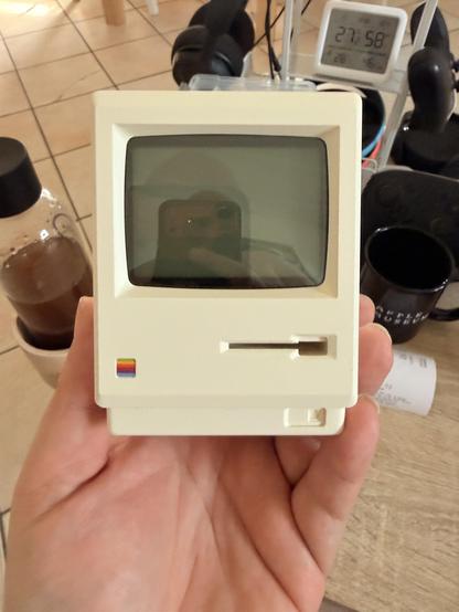 Eine Person hält einen alten Apple Macintosh Portable Computer in der Hand. Im Hintergrund sind ein Telefon und eine Kaffeemaschine.