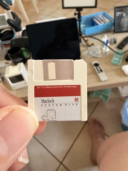 Eine Person hält eine alte Macintosh Systemdiskette vor einem Laptop auf einem Tisch mit verschiedenen elektronischen Gegenständen.