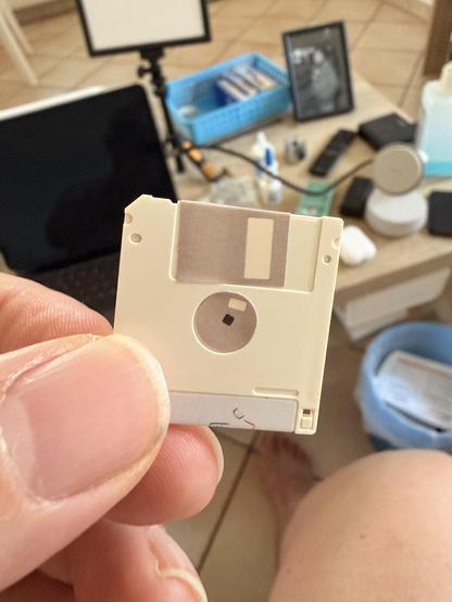 Eine Person hält eine 3,5-Zoll-Diskette vor einem Tisch mit elektronischen Geräten und Werkzeugen.