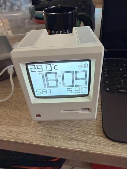 Weißer Retro-Wecker mit blauem Display auf einem Holztisch, neben einem Laptop und einer schwarzen Tasse.