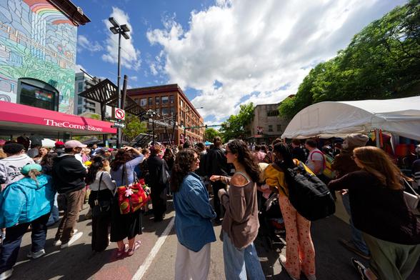 Two woman face each other and are seen side on at the bottom middle of the images and they are surrounded by a crowd that fills a canyon between two rows of buildings with cumulus clouds and blue sky above