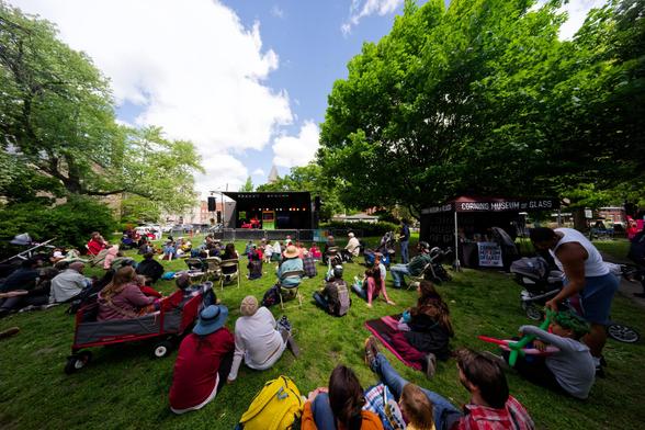 Many people sit on the grass of a large lawn in a park with trees looking towards a tiny-looking stage with a puppet stage erected inside it