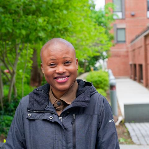Black woman wearing grey jacket smiles in front of trees and sidewalk