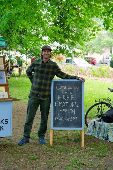 Man with flannel shirt and billed cap has arm over sign that say Come on by for FREE EMOTIONAL HEALTH Worksheet in a green park