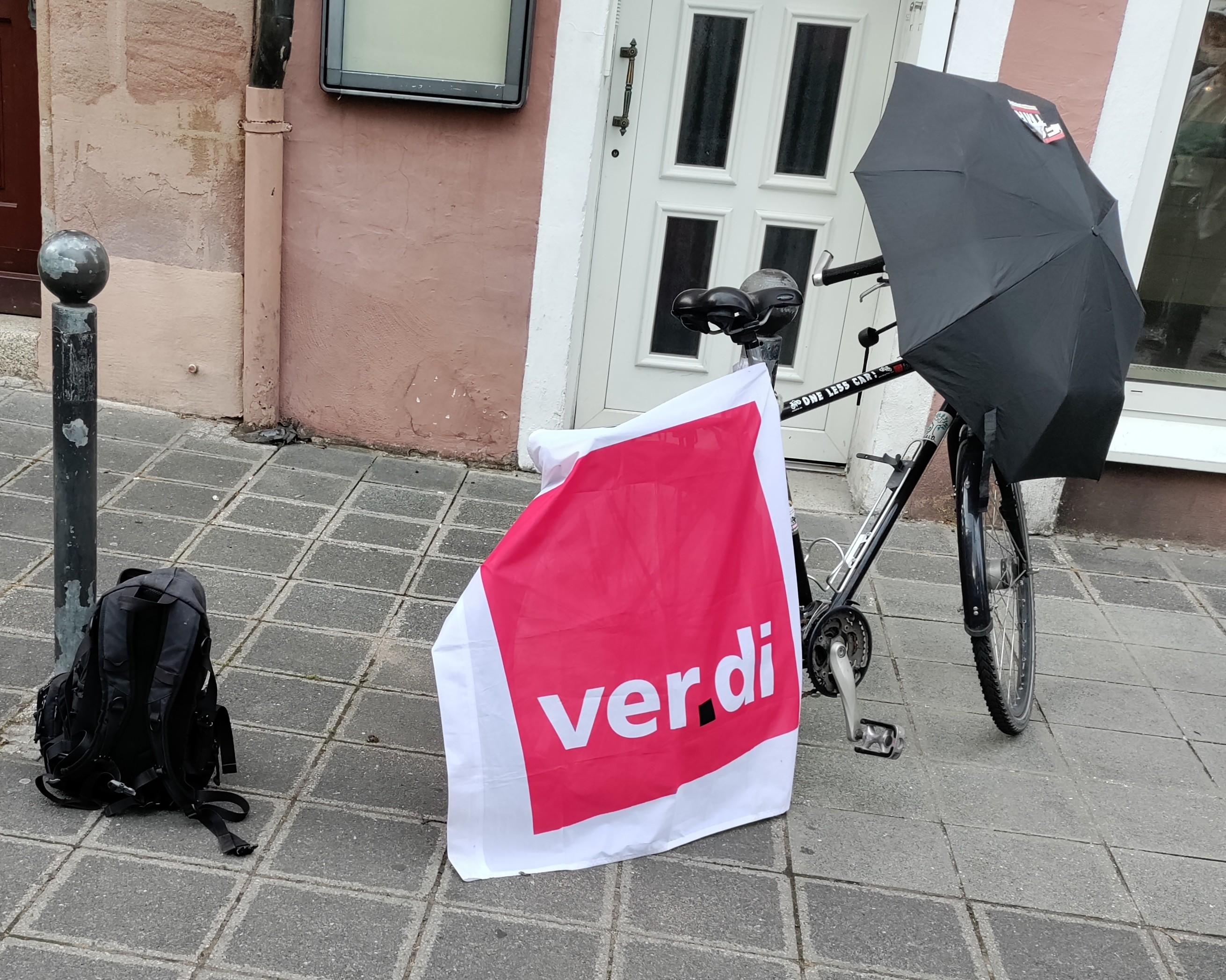 Eine Verdi Fahne hängt über ein Rad, daneben steht ein Rucksack. Über dem Steuer des Rads liegt ein geöffneter Regenschirm mit Sticker (FCK TMR - im Bild aber nicht lesbar)
