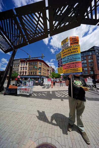 A man with a shirt that says KEEP HOPTIMISTIC with a frog graphic has a stack of signs covering his face, above him is the large metal decorative structure at the end of the commons and below that a table with a sign that says indivisible tompkins and a street buildings and more people behind that