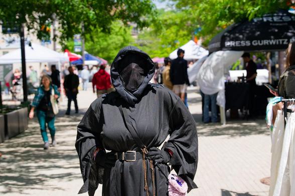 Person wears all black costume like a ninja or something on an expanse of white pavement with people and booths surrounding
