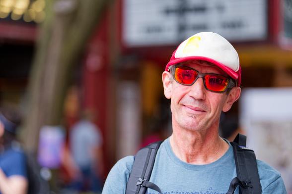 man will billed cap and orange tinted glasses wears t-shirt with backpack straps there is a theater marquee way in the back