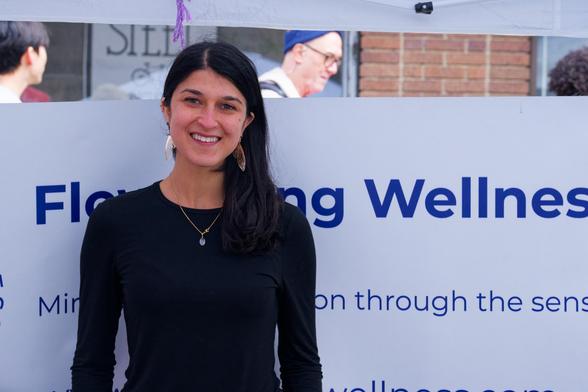 A woman wearing a blue blouse with her hair parted to one side stands in front of a sign
