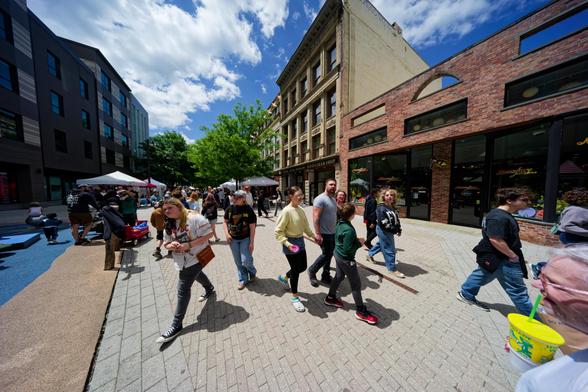 People seem to mill in all directions in a aplaza surrounded by short and moderately tall buildings on both sides under cumulus clouds.