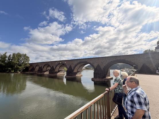 Drei Personen stehen auf einer Brücke über einem Fluss und blicken auf eine steinerne Bogenbrücke mit Wolkenhimmel.