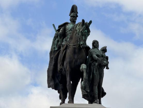 Bronzestatue eines Mannes auf einem Pferd mit zwei Frauen, unter einem blauen Himmel mit Wolken.
