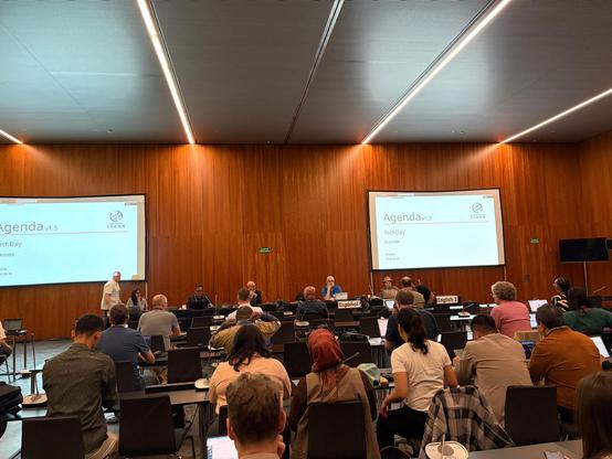 Photo from the back of a large conference room. There are rows of tables with chairs behind them where people are sitting. There is a row of tables in the front where several people sit facing the crowd. There are two large screens on the front wall that have a slide titled “Agenda 1.5”