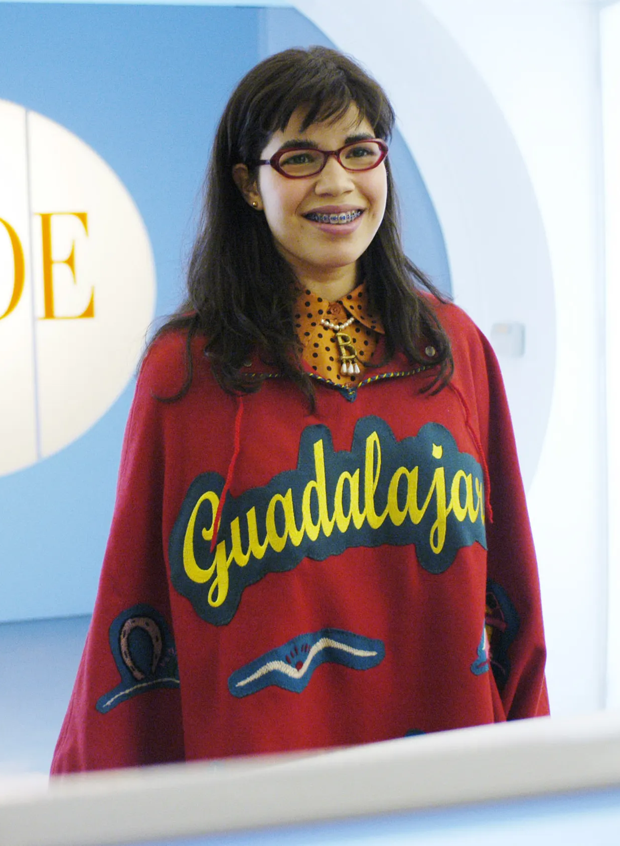 Photo of actress America Ferrera on the set of the TV show Ugly Betty. She's sporting a red "Guadalajara" poncho, wit orange/black polka-dot blouse. She's also wearing a pearl necklace with a gold "B" pendant with three teardrop pearl danglers.